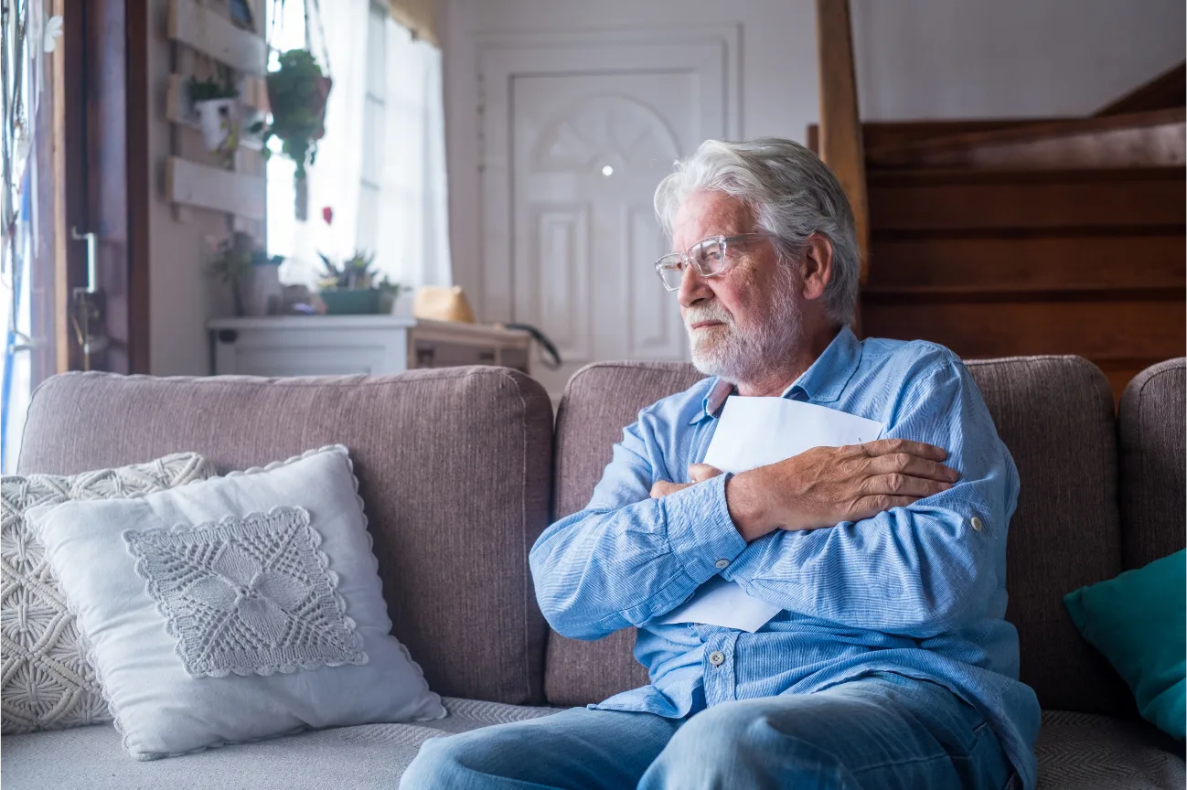 An older man sitting on a sofa at home, holding papers and looking out a window.
