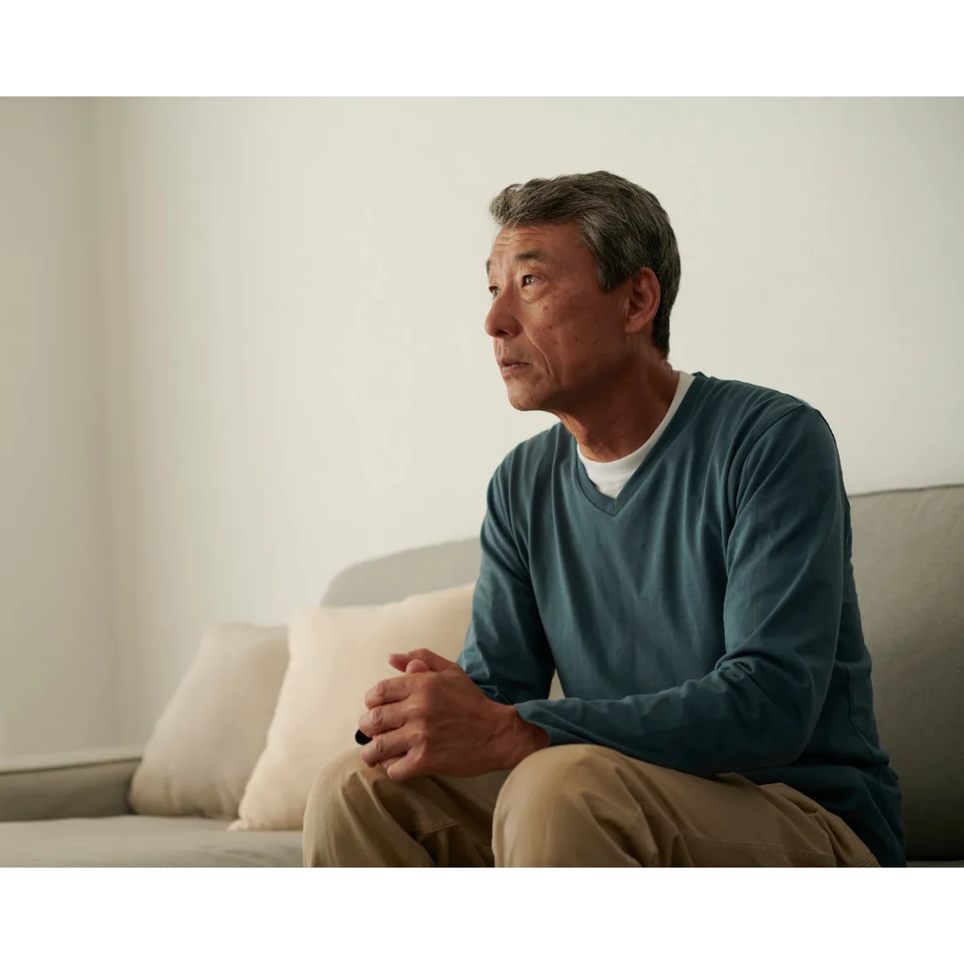 A man sitting alone on a couch at home, looking away pensively.